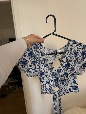 Blommig omlottopp med knyt - Söt kortärmad topp med blått blommönster på vit botten. Omlottmodell med knyt framtill och öppen detalj vid ryggen. Luftigt material och volangiga ärmar ger en romantisk vibe. Perfekt för varma dagar och trendiga outfits.