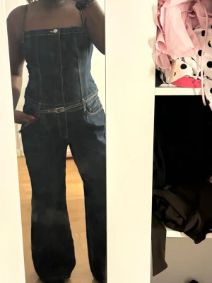 Mörkblå denim jumpsuit med vida ben - Snygg mörkblå denim jumpsuit med raka, vida ben och smala axelband. Har markerad midja med bälte och vita kontrastsömmar. Klassisk knappdetalj framtill och fickor på sidorna. Perfekt för en trendig och avslappnad look.