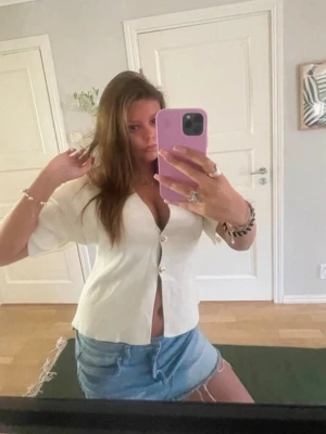 Vit kortärmad blus med knappar - Trendig vit blus med korta ärmar och djup v-ringning. Blusen har en figurnära passform och stängs med stora dekorativa knappar framtill. Perfekt att matcha med jeans eller kjol för en snygg look.