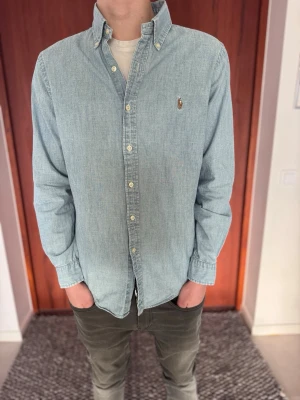 Ralph Lauren Denimskjorta – Ljusblå (Slim Fit) - Säljer en snygg ljusblå denimskjorta (chambray) från Polo Ralph Lauren med marinblå logga. Det är en Slim Fit-modell som är något liten i storleken, så den sitter mer som en Small än en Medium. • Märke: Polo Ralph Lauren • Storlek: Medium men passar även Small • Passform: Slim Fit • Skick: Bra begagnat skick utan några slitage • Nypris: ca 1700kr