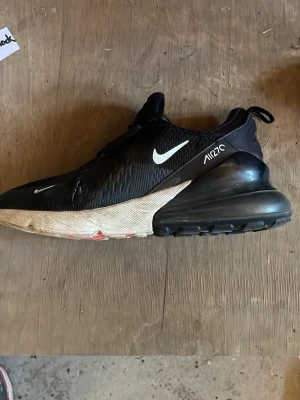 Nike Air Max 270 svart/vit sneaker - Nike Air Max 270 sneakers i svart med vit sula och tydlig Air Max-dämpning i hälen. Skon har en sportig siluett, mesh-överdel och vita detaljer som Swoosh-loggan och texten 'AIR270' på sidan. Perfekt för dig som gillar streetwear och bekväma sneakers.