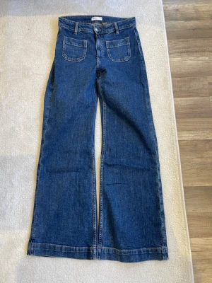 Blåa vida jeans med fickor fram - Säljer ett par blåa jeans med hög midja och raka, vida ben. Jeansen har två stora framfickor och bälteshällor. Klassisk blå denim med synliga sömmar. Perfekta för en avslappnad och trendig stil.