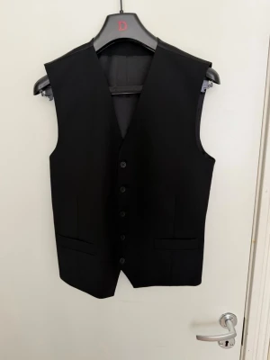 Svart kostymväst från Dressmann XS - Klassisk svart kostymväst från Dressmann i storlek XS. Västen har fem knappar framtill, två fickor och är tillverkad i ett slätt tyg med blankt foder. Perfekt för en stilren och uppklädd look.