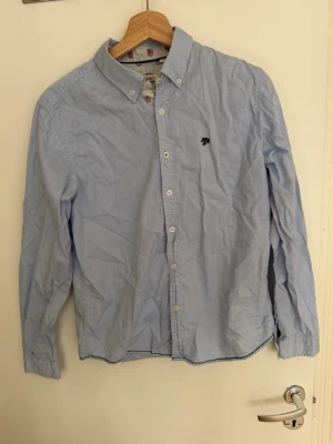 Ljusblå skjorta Hampton Republic - Klassisk ljusblå skjorta från Hampton Republic med button-down krage och broderad logga på bröstet. Skjortan har vita knappar och tunna ränder på insidan av kragen samt röda emblem. Tillverkad i bomull för en bekväm känsla.