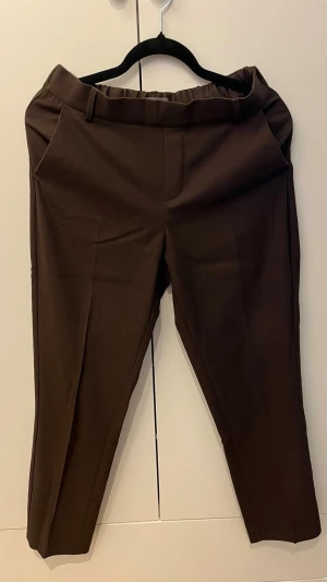 Bruna kostymbyxor - Snygga bruna kostymbyxor med rak passform och resår i midjan. Byxorna har pressveck, bälteshällor och två sidofickor fram samt två passpoalfickor bak. Perfekta för en clean och stilren look.