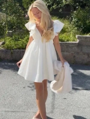 Vit kortklänning med volang och rosett - Supersöt vit kortklänning med stora volangärmar och v-ringning med snörning framtill. Klänningen har en stor rosett i ryggen och är tillverkad i ett lätt bomullstyg, perfekt för varma dagar. Fladdrig och luftig passform som ger en romantisk vibe, Den är helt ny har aldrig använt bara testat en gång men va lite för stor för mig. Kom privat för fler bilder ifall det behövs💝