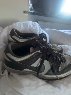 Nike Dunk Low svart och grå sneakers - Säljer ett par Nike Dunk Low sneakers i svart och grått med vita detaljer. Klassisk låg modell med snörning och ikoniska swoosh-loggan på sidan. Skorna har en sportig vibe och passar perfekt till streetwear-stilen.
