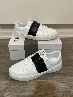 Valentino skor - Snygga vita sneakers från Valentino Garavani med en bred svart rem över vristen dekorerad med nitar. Skorna har snörning och är tillverkade i skinn för en lyxig känsla. Perfekta för dig som vill ha en clean men ändå edgy stil.