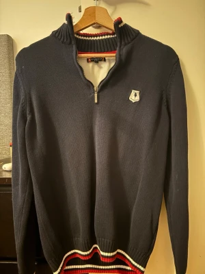 Mörkblå stickad tröja från Polo - Mörkblå stickad tröja från Polo med halv dragkedja och hög krage. Tröjan har vita och röda detaljer vid krage och nederkant samt ett broderat Polo-märke på bröstet. Perfekt för dig som gillar klassisk och sportig stil.