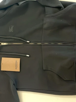 Svart hoodie med dragkedja Burberry - Snygg svart hoodie från Burberry med dragkedja framtill, huva med dragsko och två stora fickor. Tillverkad i mjukt material och har diskret Burberry-logga på bröstet. Perfekt för en clean och stilren look.