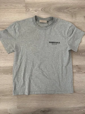 Grå Essentials Fear of God t-shirt - Säljer en stilren grå t-shirt från Essentials Fear of God. T-shirten har korta ärmar, rund halsringning och en liten svart logga på bröstet. Tillverkad i mjuk bomull för maximal komfort och enkel att matcha med allt. Storlek S