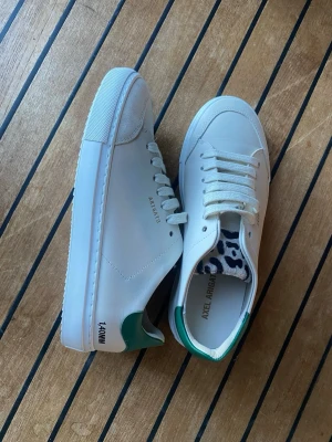 Vita Axel Arigato sneakers med grönt - Säljer ett par stilrena vita sneakers från Axel Arigato med gröna detaljer på hälen. Skorna har klassisk låg profil, vita snören och ovandel i slätt läder. Perfekta för dig som gillar minimalistisk och modern design.