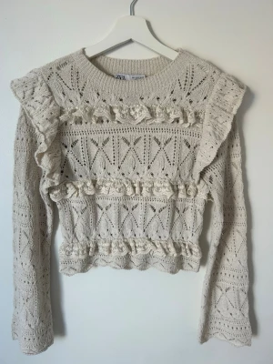 Vit spetsstickad tröja från Zara - Säljer en vit spetsstickad tröja från Zara med dekorativa volanger och virkade blommor framtill. Tröjan har lång ärm, rund halsringning och ett detaljerat hålmönster över hela plagget. Perfekt för dig som gillar romantisk och bohemisk stil.