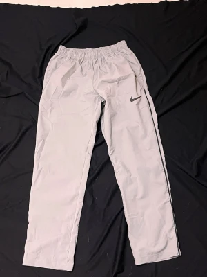 Gråa träningsbyxor från Nike - Snygga gråa träningsbyxor från Nike med Dri-FIT-material som håller dig torr under träningen. Byxorna har elastisk midja och en diskret Nike-logga på vänster ben. Perfekta för gymmet eller löprundan.