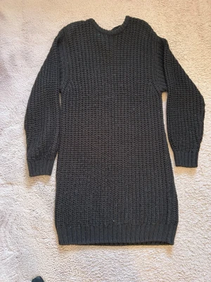 Svart stickad klänning med knyt - Mysig svart stickad klänning med lång ärm och djup v-ringning i ryggen. Knytband i nacken ger en snygg detalj och extra stil. Perfekt för kyligare dagar och enkel att matcha med boots eller sneakers.