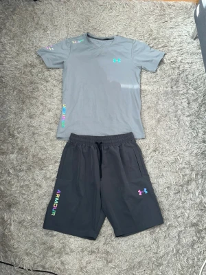 Under Armour set i grått (reflex) - Snyggt träningsset från Under Armour med en ljusgrå t-shirt och mörkgrå shorts. T-shirten har färgglada loggor och detaljer på ärmen och sidan, medan shortsen har elastisk midja, dragsko och färgglad logga samt texten 'ARMOUR' på benet. 