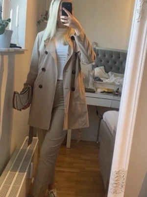 Beige trenchcoat med knappar - Stilren beige trenchcoat med dubbelknäppning och klassisk krage. Jackan har stora bruna knappar och raka ärmar.  Slits baktill med ett skärp i midjan. Bra skick.