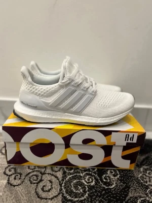 Vita Adidas Ultra Boost sneakers - Säljer ett par helvita Adidas Ultra Boost sneakers med stickad ovandel i mesh och klassiska tre ränder på sidan. Skorna har en bekväm Boost-sula och vita snören. Perfekta för dig som gillar stilrena och sportiga sneakers.