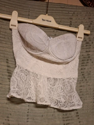 Vit spetskorsett med volang - Supersöt vit korsettopp från Bik Bok i spets med vadderade kupor och volang nedtill. Toppen har dragkedja i ryggen och är perfekt för dig som vill ha en feminin och trendig look. Spetsdetaljerna ger en romantisk vibe och toppen är axelbandslös.
