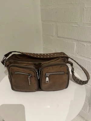 Brun axelväska från Noella - Cool brun axelväska i mockaimitation från Noella med två stora framfickor med dragkedja och en bred axelrem dekorerad med silvriga nitar. Perfekt för dig som gillar en lite tuffare stil och vill ha plats för allt du behöver.