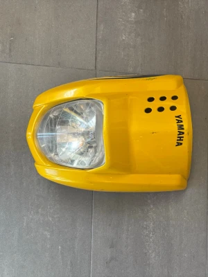 Gul frontkåpa med strålkastare Yamaha - Säljer en gul frontkåpa med inbyggd strålkastare från Yamaha. Kåpan har flera ventilationshål och tydlig Yamaha-logga på sidan. Passar till moped eller motorcykel och är tillverkad i plast.