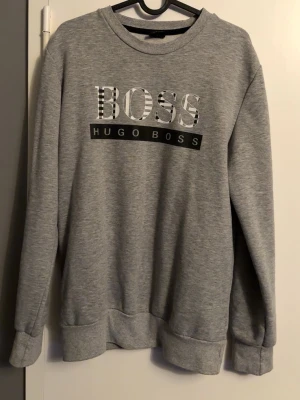 Grå sweatshirt från Hugo Boss - Snygg grå sweatshirt från Hugo Boss med stort BOSS-tryck och logga på bröstet. Tröjan har rund halsringning, långa ärmar och ribbade muddar vid ärmslut och nederkant. Perfekt för en avslappnad och stilren look.