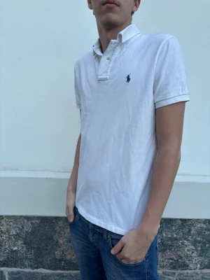 Ralph Lauren pike - Klassisk vit pikétröja från Ralph Lauren med korta ärmar och den ikoniska loggan broderad på bröstet. Tröjan har krage och knappar vid halsen. Perfekt till jeans eller shorts för en clean och stilren look.