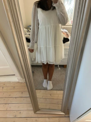 Vit prickig kortklänning med volang - Söt vit kortklänning med diskret prickigt mönster, volangdetaljer och v-ringning. Klänningen har långa ärmar med smock vid ärmslut och lätt rynkad kjol. Perfekt för sommarens alla tillfällen.