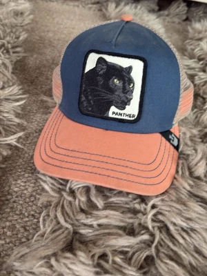 Panther keps från Goorin bros - Säljer en snygg truckerkeps från Goorin Bros med blå framsida, orange skärm och mesh i beige. På framsidan finns ett broderat märke med en svart panter och texten 'Panther'. Justerbar snapback baktill för perfekt passform.