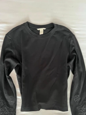 Svart långärmad topp med spetsdetaljer - Snygg svart långärmad topp från H&M med unika spetsdetaljer på ärmarna. Toppen har en klassisk rund halsringning och är gjord i mjuk bomull som känns skön mot huden. Perfekt för dig som vill ha en enkel men ändå speciell topp.