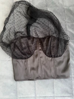 Svart korsettopp one-shoulder - Trendig svart korsettopp med transparent mesh över axeln och prickigt mönster. Toppen har markerade kupor och dragkedja bak. Perfekt för dig som vill sticka ut med en edgy och cool look.