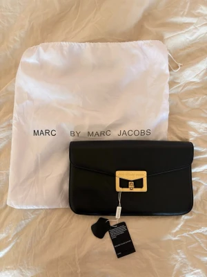 Svart kuvertväska Marc by Marc Jacobs - Svart kuvertväska från Marc by Marc Jacobs i äkta italienskt läder. Väskan har en rektangulär form och stängs med ett guldfärgat metallspänne med logan. Den ör aldrig använd och har prislappen kvar. 