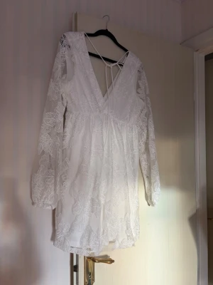 Vit spetsklänning med v-ringning - Kort vit klänning med djup v-ringning och långa ärmar i transparent spets. Klänningen har fina broderade blommor, knytband i nacken och en luftig passform. 