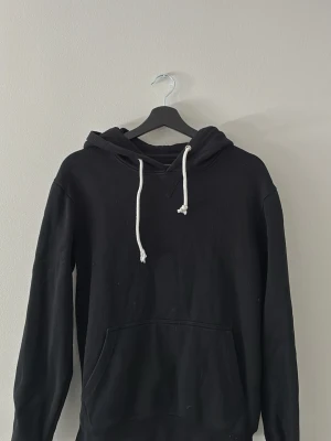 Svart hoodie med vita snören - En klassisk svart hoodie med vita dragsnören i huvan och stor magficka framtill. Hoodien har en avslappnad passform och är tillverkad i mjukt bomullsmaterial. Perfekt för en chill och enkel stil.