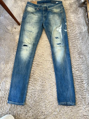 Dondup George - Säljer ett par blå skinny jeans med slitningar och hål på framsidan. Jeansen har en tvättad look med ljusare partier och klassisk femficksmodell. Perfekta för dig som gillar en avslappnad och trendig stil. Storlek 32, väldigt fina. 