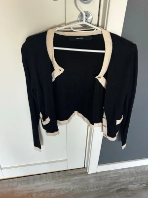 Svart kofta med beige detaljer - Stilren svart kofta från Vero Moda med beige kontrastkanter vid krage, knappslå, fickor, ärmslut och nederkant. Koftan har rund halsringning, dekorativa fickor framtill och guldknappar. Perfekt för en chic och enkel look.