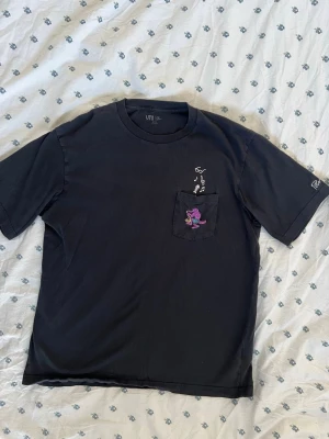 Svart Uniqlo t-shirt med ficka - Svart limited edition Fergadelic purple crocodile / lila krokodil t-shirt från Uniqlo x Fergus Purcell med en bröstficka som har ett broderat motiv av en lila krokodil och musiknoter. T-shirten har rund halsringning och korta ärmar. Perfekt för dig som gillar coola detaljer och streetwear vibes. Storlek L / large. Måtten finns. 