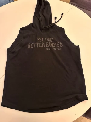 hoodie från Better Bodies - Svart ärmlös hoodie från Better Bodies med tryck 'EST. 1982 BETTER BODIES NEW YORK CITY' på bröstet. Huva med dragsko och avslappnad passform, perfekt för gymmet eller träning. Materialet är mjukt och stretchigt.