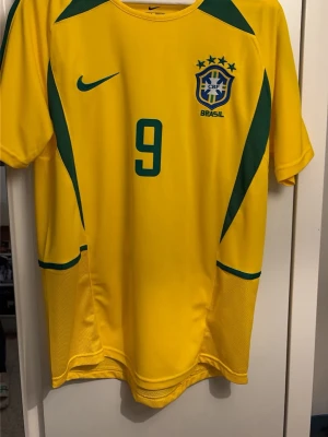 Brasilien fotbollströja Nike nr 9 - Gul Brasilien fotbollströja från Nike med gröna detaljer och nummer 9 på bröstet och ryggen. Tröjan har korta ärmar, rund hals och är tillverkad i ett lätt syntetmaterial som andas. Perfekt för fotbollsträning eller att bära som supporter.
