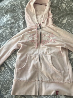 Ljusrosa Ed Hardy hoodie med dragkedja - Säljer en ljusrosa hoodie från Ed Hardy i mjuk velour med dragkedja framtill och broderad logga i vitt och rosa på bröstet. Hoodien har fickor fram, huva med dragsko och dekorativa sömmar längs nederkanten. Perfekt för en chill och trendig look.