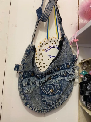 Jeansväska med fransar och nitar - Cool handväska i blått denim med slitna fransdetaljer och nitar framtill. Väskan har en justerbar axelrem och en dekorativ ficka på framsidan. Perfekt för dig som gillar unik och retro stil.