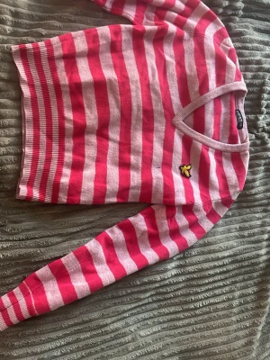 Rosa-randig stickad tröja Lyle & Scott - Stickad tröja med breda rosa och ljusrosa ränder från Lyle & Scott. Tröjan har V-ringning, långa ärmar och en gul broderad fågel på bröstet. Ribbstickade muddar vid ärmslut och nederkant. Perfekt för dig som gillar färg och mönster.