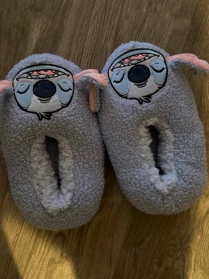 Grå Stitch-tofflor i fleece - Mysiga grå tofflor med Stitch-motiv från Disney. Tillverkade i mjuk fleece med broderat ansikte och rosa detaljer på öronen. Perfekta för att hålla fötterna varma hemma och har en gullig design som sticker ut.
