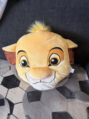 Simba kudde från Lejonkungen - Mjuk och gosig kudde formad som Simbas ansikte från Lejonkungen. Kudden är gul med bruna och vita detaljer samt en liten tofs av gult fluff på toppen. Perfekt som prydnad eller mysig kompis i soffan.