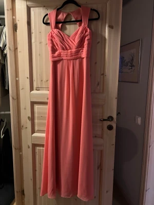 Korall balklänning  - Säljer en elegant långklänning i rosa med breda axelband och v-ringad omlott framtill. Klänningen har ett markerat midjeband och ett flytande, svepande fall. Perfekt för speciella tillfällen och ger en romantisk look.