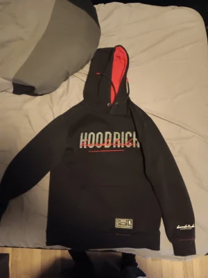 Svart HOODRICH hoodie med röd detalj - Snygg svart hoodie från Hoodrich med stor logga på bröstet och ryggen. Huvan har röd insida och dragsko. Detaljerad patch nertill och broderad text på ärmen. Perfekt för dig som gillar streetwear och vill sticka ut med coola detaljer.