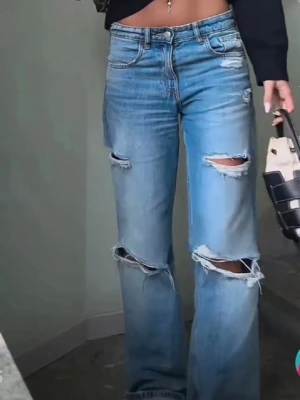 Blå wide jeans med slitningar - Säljer ett par blå jeans med breda ben och snygga slitningar framtill. Klassisk femficksmodell med knapp och dragkedja i midjan. Jeansen har en cool, avslappnad vibe och är gjorda i mjukt denimtyg. Perfekta för dig som gillar streetstyle och vill sticka ut.