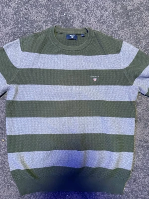 Stickad GANT-tröja i bomull – Storlek M – Fint skick - Säljer en riktigt snygg stickad tröja från GANT i en härlig olivgrön och grå färgkombination. Tröjan har en snygg struktur i tyget och den klassiska loggan på bröstet. Perfekt för både vardag och lite finare tillfällen. • Märke: GANT • Storlek: M • Skick: Mycket bra • Material: Mjuk och skön bomull Passa på att fynda en tidlös klassiker till garderoben! Fraktar snabbt. 