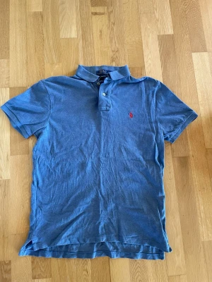 Blå pikétröja från Polo Ralph Lauren - Snygg blå pikétröja från Polo Ralph Lauren med klassisk krage, två knappar och röd broderad logga på bröstet. Tillverkad i mjuk bomull och har korta ärmar för en chill look. 
