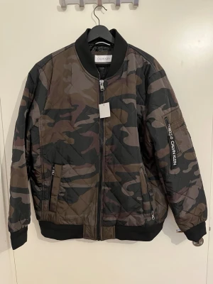 Camouflage bomberjacka Calvin Klein - Snygg bomberjacka från Calvin Klein i camouflage-mönster med svarta ribbade muddar och dragkedja framtill. Jackan har fickor med dragkedja och en extra ficka på ärmen med Calvin Klein-logga. Fodrad insida med diskret logomönster. Aldrig använts.
